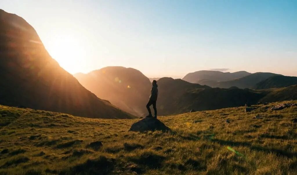 Silhouettenfotografie einer Person, die während der goldenen Stunde auf grünem Gras vor Bergen steht