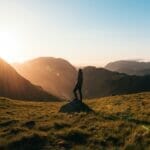 silhouette photography of person standing on green grass in front of mountains during golden hour