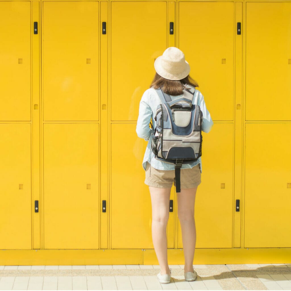 A woman with a backpack is standing in front of yellow lockers.
