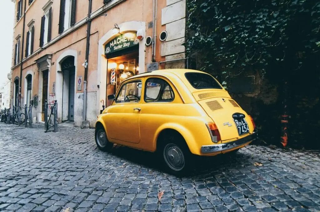 A yellow fiat 500 is parked on a cobblestone street on its journey from Perth to Rome.