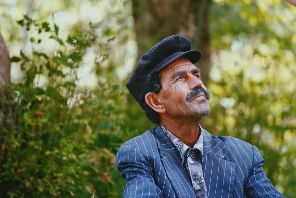 An old man with Balkan facial features sitting on a bench in the woods.