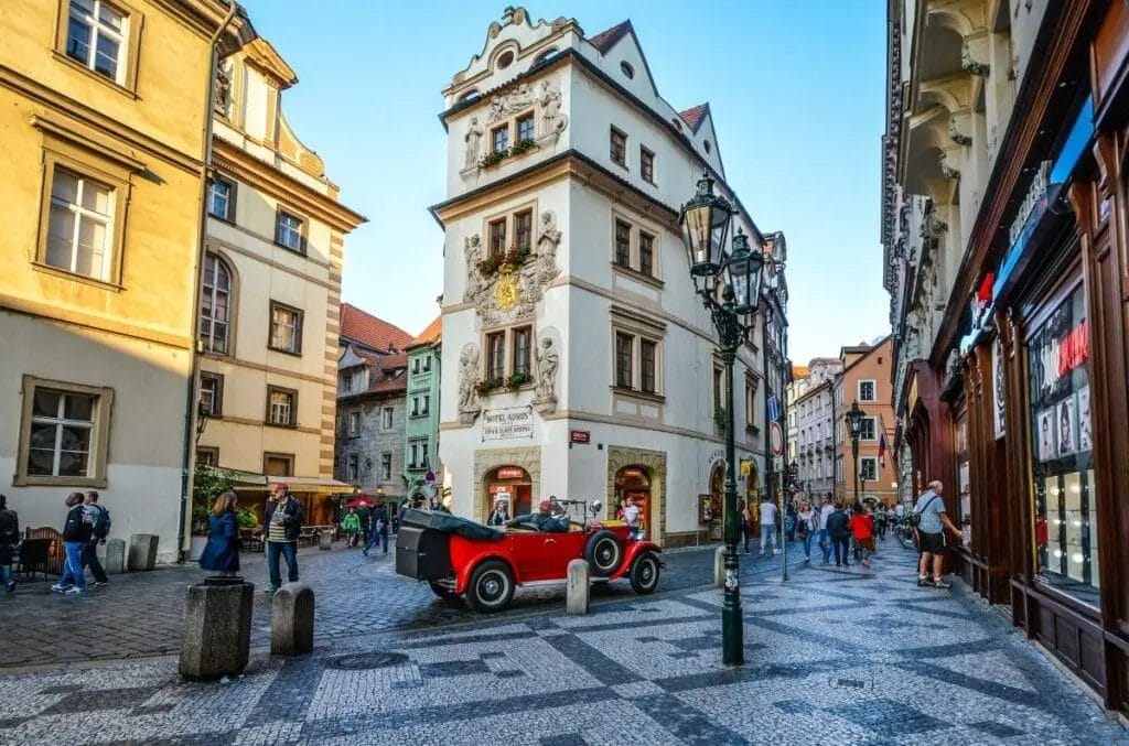 A vintage red convertible car parked on a cobblestone street in a historic European city bustling with pedestrians and showcasing European fashion do's and don'ts.