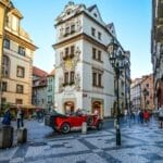 A vintage red convertible car parked on a cobblestone street in a historic European city bustling with pedestrians and showcasing European fashion do's and don'ts.