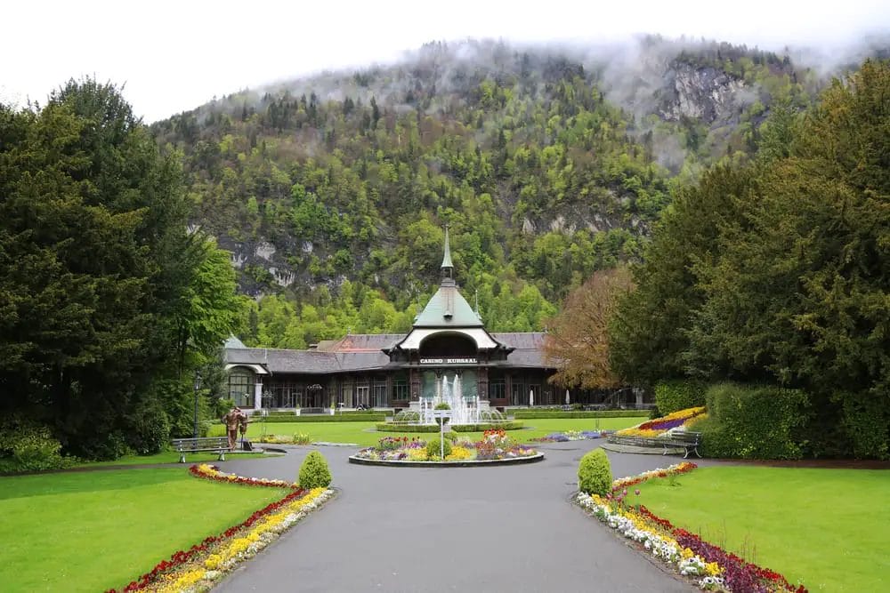 A well-manicured garden leading up to a traditional building with a green roof against a backdrop of mountains partially shrouded in mist.