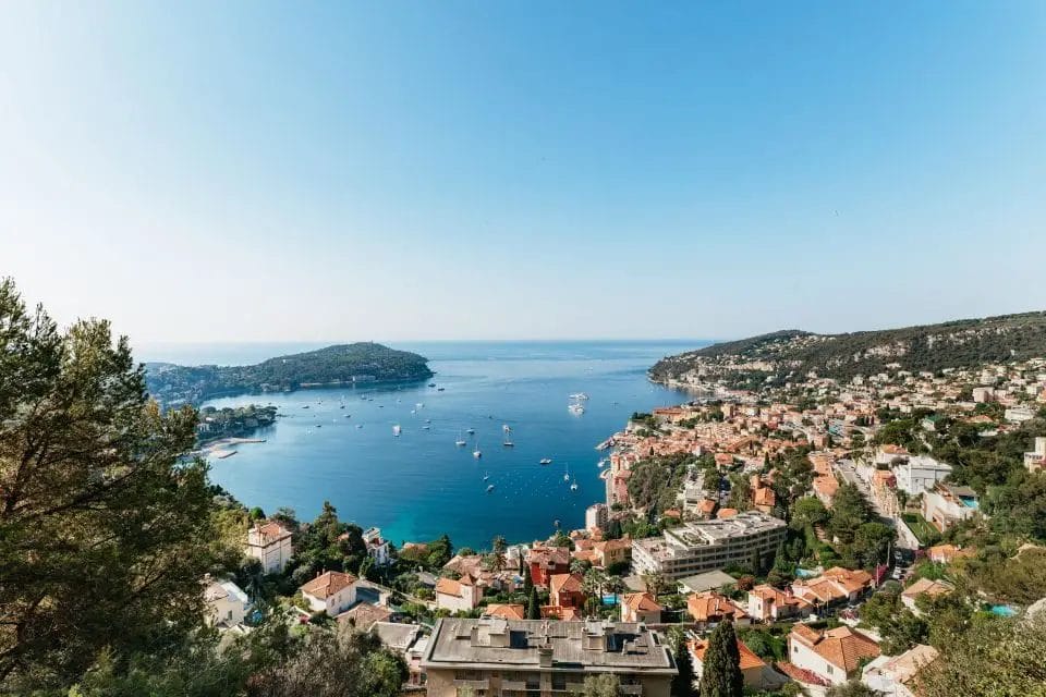 A panoramic view of a coastal town with boats on clear blue waters, surrounded by green hills under a blue sky, perfectly capturing the essence of a day trip to Villefranche-Sur-Mer from